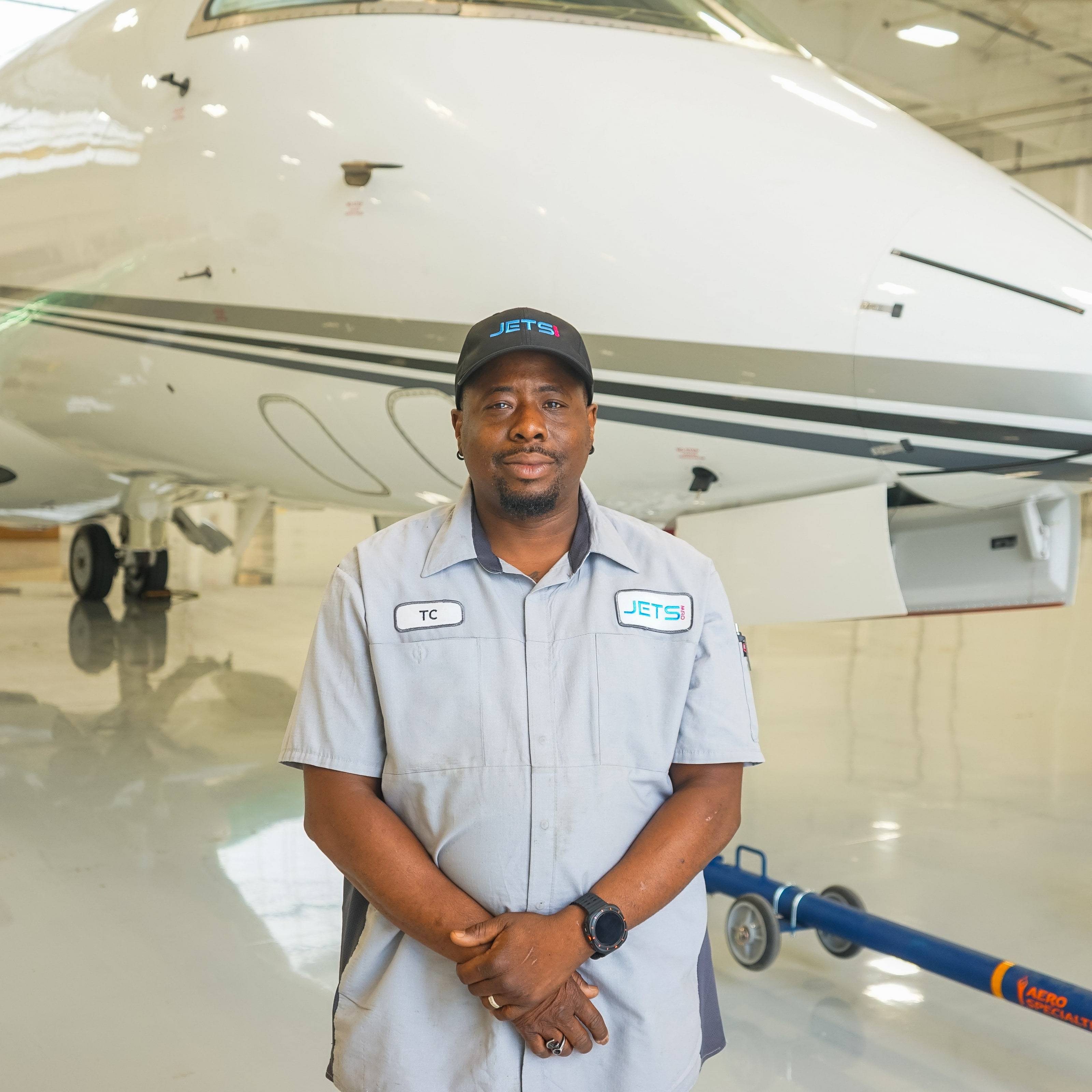 Jets MRO technician in a grey uniform shirt and cap standing in front of a business jet in the hangar